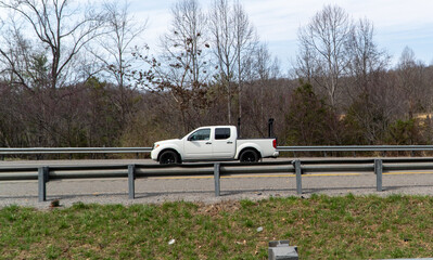 White pickup truck with a black utility rack driving on a highway, flanked by a metal guardrail and a background of bare trees under a bright sky, symbolizing transportation and journey. © Michael