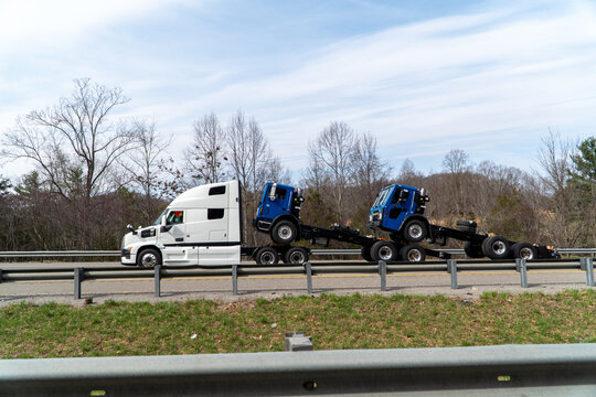 White semi-truck transporting two new blue Mack truck chassis on a highway under a clear sky, showcasing heavy haul logistics and commercial vehicle delivery.