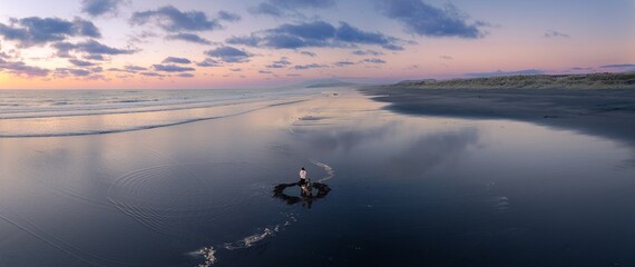 A couple stands on the black sands of Kawhia hotwater Beach, New Zealand, at sunset. They are digging in the sand, creating a small pool of water. The sky is pink and purple. © Stefan