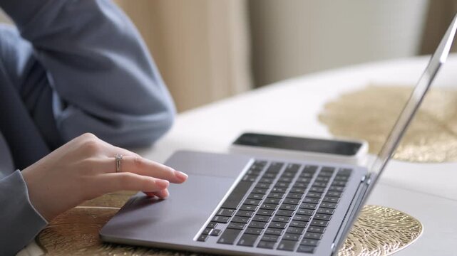 Person uses laptop on table. Hand rests on keyboard. Phone lies nearby. Golden textured mat under device