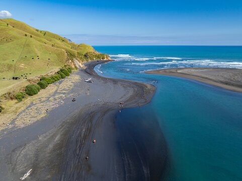 Aerial view of people enjoying a sunny day at the black sand beach of Marokopa, New Zealand. Vehicles are parked nearby.