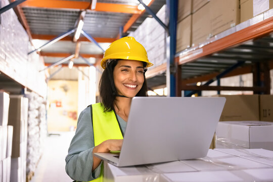 Smiling Latina female worker in safety vest and hardhat using laptop in industrial warehouse, perform logistics operations, managing stock supply chain, checking inventory in distribution center