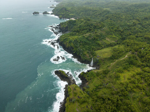 Aerial view of Banyu Tibo Waterfall cascading into the ocean along the rocky coastline with lush green terraced hills, Girisubo, Special Region of Yogyakarta, Indonesia.