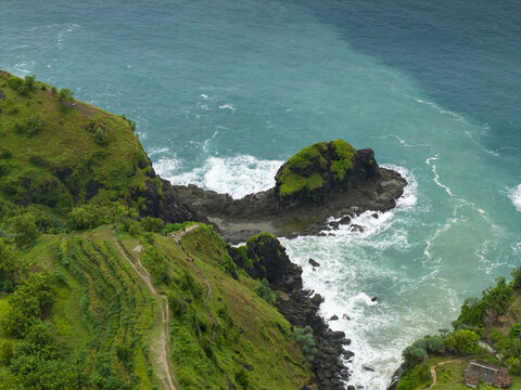 Aerial view of Banyu Tibo Waterfall cascading onto the rocky shore with turquoise ocean waves and lush green cliffs in Girisubo, Special Region of Yogyakarta, Indonesia.