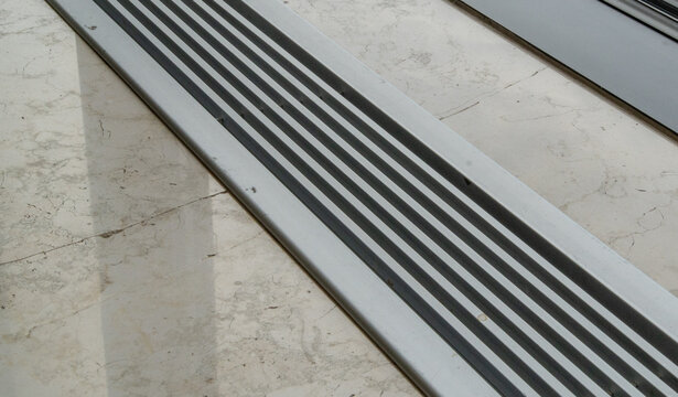 Close-up of a textured anti-slip or drainage strip with parallel lines on a worn, cracked marble or concrete floor, creating a stark geometric pattern with contrasting shadows.
