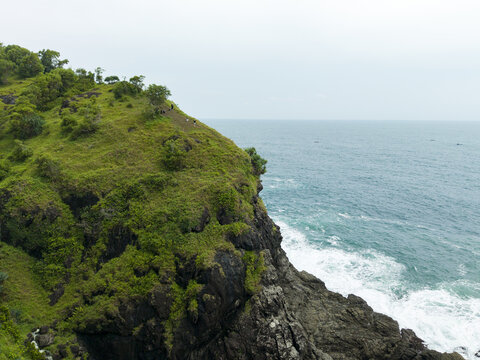 Aerial view of the lush green cliffs at Banyu Tibo Waterfall with waves crashing against the rocky shoreline in Girisubo, Special Region of Yogyakarta, Indonesia.
