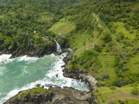 Aerial view of Banyu Tibo Waterfall cascading into the turquoise ocean waves surrounded by lush green cliffs and terraced hills in Girisubo, Special Region of Yogyakarta, Indonesia.