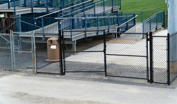 Secure chain-link fencing and dark gates provide access to empty blue stadium bleachers with railings overlooking a pristine green sports field, symbolizing venue closure or pre-event quiet.