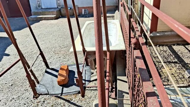 Outdoor bathtub rests on gravel beneath red metal frame with swing seat and brick, capturing unusual rustic setup blending repurposed materials in a creative yard scene