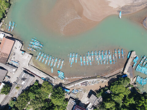 Aerial view of traditional blue fishing boats moored along the concrete pier at Baron Beach with turquoise water and sandy shores Tanjungsari, Special Region of Yogyakarta, Indonesia.