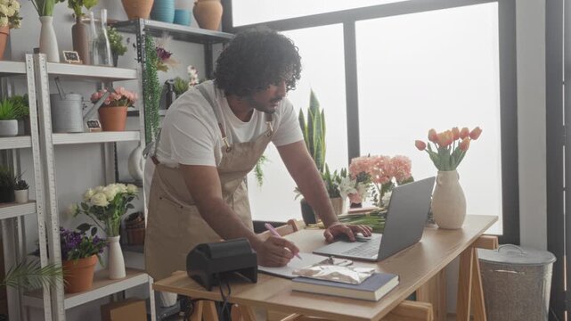 Man florist in apron leans over table typing on laptop and writes with pen in building; calm concentration.