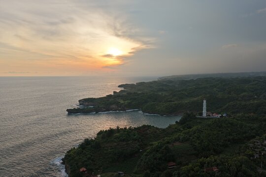 Aerial view of Baron Beach featuring the white lighthouse on rugged cliffs overlooking the Indian Ocean during a golden sunset in Tanjungsari, Special Region of Yogyakarta, Indonesia.