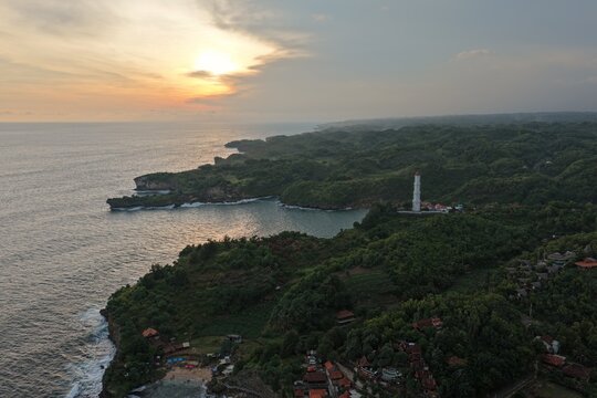 Aerial view of Baron Beach featuring the white lighthouse on a cliff overlooking the rugged coastline and sunset in Tanjungsari, Special Region of Yogyakarta, Indonesia.