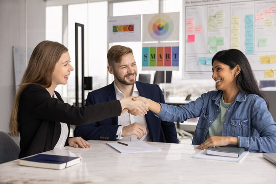 Three professionals take part in negotiations in modern conference room, two women shaking hands, finalize successful meeting, making deal, conclude hiring process seated at desk. Business, etiquette