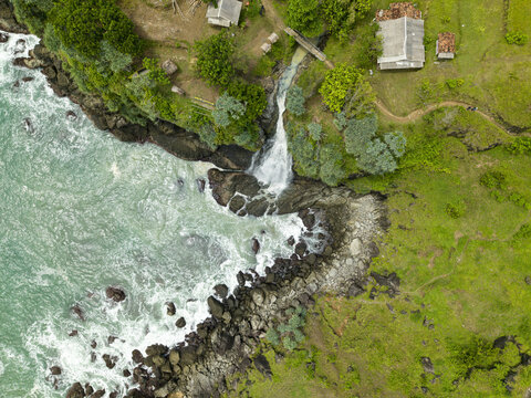 Aerial view of Banyutibo Water Fall cascading over rocky cliffs into the turquoise ocean waves near lush green hills and rural houses in Girisubo, Special Region of Yogyakarta, Indonesia.