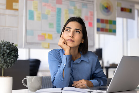Thoughtful Latina businesswoman sit at office desk with laptop, coffee mug and notebook, taking break during workday to think on business idea, reflects on work task, consider decisions and planning