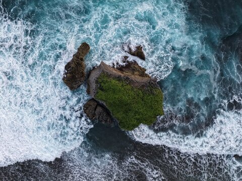 Aerial view of Baron Beach rocky outcrop with green vegetation surrounded by crashing waves and turquoise ocean water Tanjungsari, Special Region of Yogyakarta, Indonesia.
