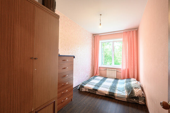 compact bedroom with single bed under window with peach curtains, wooden wardrobe, chest of drawers, dark flooring, light pink walls, simple pendant light. Natural light fills space
