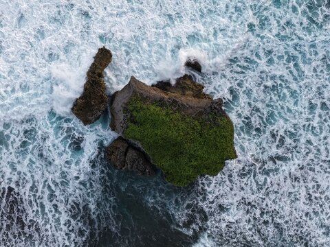 Aerial view of a rocky outcrop covered in green moss surrounded by crashing white waves and turquoise ocean water at Baron Beach in Tanjungsari, Special Region of Yogyakarta, Indonesia.