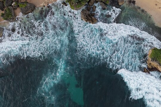 Aerial view of Baron Beach with turquoise waves crashing against dark volcanic rocks and a sandy shoreline Tanjungsari, Special Region of Yogyakarta, Indonesia.