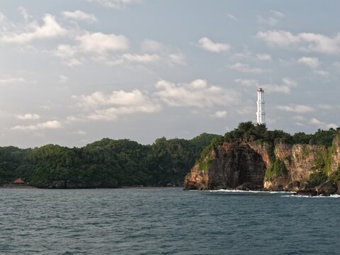 Aerial view of Baron Beach lighthouse perched on a rocky cliff overlooking the sea and lush green hills in Tanjungsari, Special Region of Yogyakarta, Indonesia.