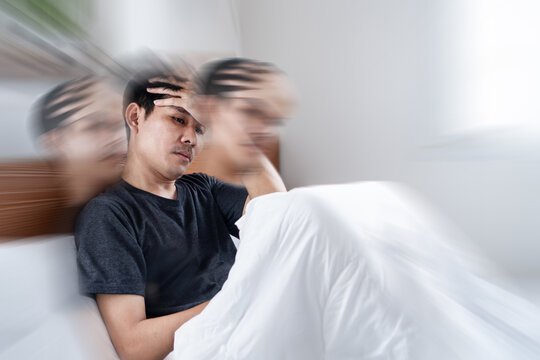 Asian man sitting on a bed feeling dizzy, suffering from a severe headache, migraine, or vertigo. The blurred motion effect illustrates his disorientation, physical pain, and mental stress.