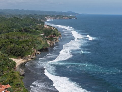 Aerial view of Baron Beach with white waves crashing against the dark shoreline and lush green cliffs in Tanjungsari, Special Region of Yogyakarta, Indonesia.