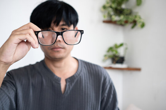 A young Asian man holding a pair of modern black-rimmed eyeglasses in front of his face. The shot highlights vision care, optometry, clear sight, and optical health concepts.