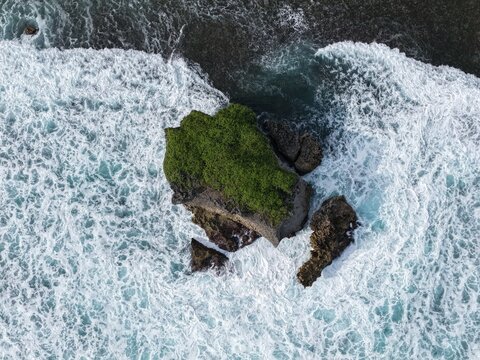 Aerial view of Baron Beach featuring a rocky island covered in green vegetation surrounded by crashing white waves and turquoise ocean water in Tanjungsari, Special Region of Yogyakarta, Indonesia.