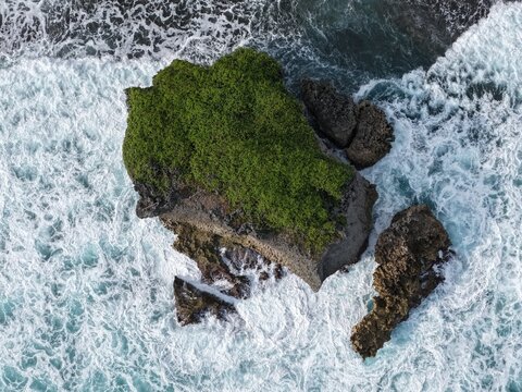 Aerial view of a lush green rocky outcrop surrounded by crashing white waves at Baron Beach in Tanjungsari, Special Region of Yogyakarta, Indonesia.