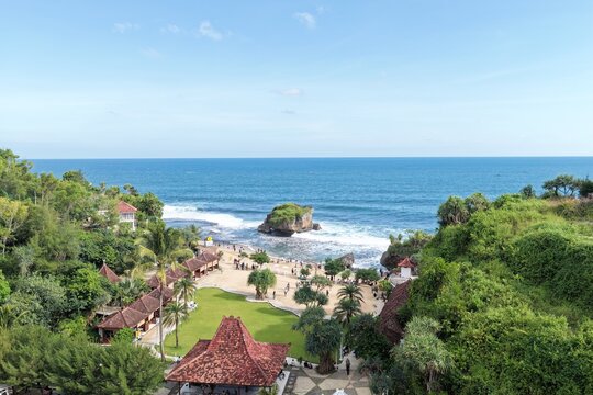 Aerial view of Baron Beach with its iconic rock formation, sandy shore with people, and traditional red-roofed buildings in lush greenery in Tanjungsari, Special Region of Yogyakarta, Indonesia.