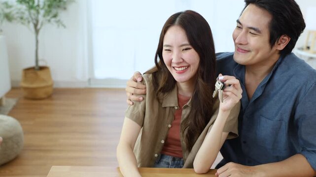 Asian man and woman sitting on sofa receiving house keys from real estate agent, representing final stage of buying process, showing joy, commitment, trust, and achievement in new home journey