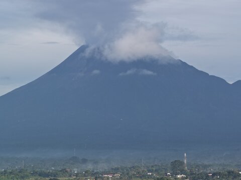 Aerial view of Mount Merapi volcano with a plume of smoke at the summit overlooking a lush landscape and distant buildings Tanjungsari, Special Region of Yogyakarta, Indonesia.