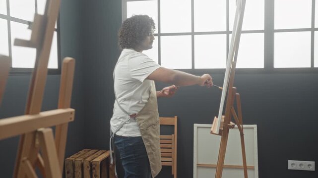 Man painter wearing apron holds paintbrush and palette while painting on easel in studio; creative concentration.