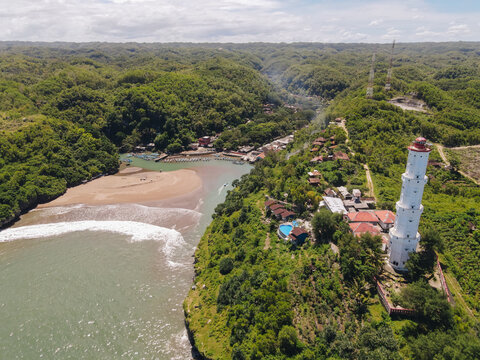 Aerial view of Baron Beach featuring the white lighthouse, sandy cove, and lush tropical hills in Tanjungsari, Special Region of Yogyakarta, Indonesia.