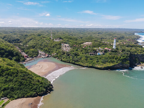 Aerial view of Baron Beach with its white lighthouse on a cliff overlooking the turquoise ocean and lush green hills in Tanjungsari, Special Region of Yogyakarta, Indonesia.