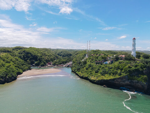 Aerial view of Baron Beach with its iconic white lighthouse standing on a lush green cliff overlooking the turquoise sea in Tanjungsari, Special Region of Yogyakarta, Indonesia.