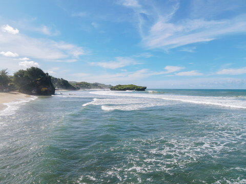 Aerial view of Baron Beach with rolling waves, sandy shoreline, and rocky cliffs under a bright blue sky in Tanjungsari, Special Region of Yogyakarta, Indonesia.