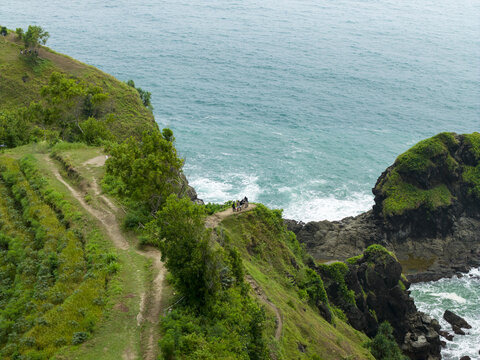 Aerial view of the lush green cliffs of Banyu Tibo Waterfall with people standing on a path overlooking the turquoise ocean in Girisubo, Special Region of Yogyakarta, Indonesia.