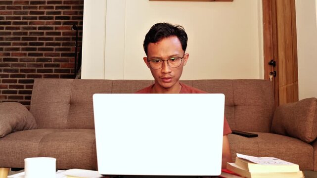 An Asian man working seriously on a laptop at home, focusing on his tasks in a comfortable indoor environment. This scene represents remote work, productivity, online business, and modern digital life