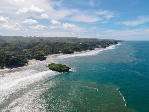 Aerial view of Baron Beach with turquoise ocean waves crashing against a rocky outcrop and lush green hills under a bright sky Tanjungsari, Special Region of Yogyakarta, Indonesia.