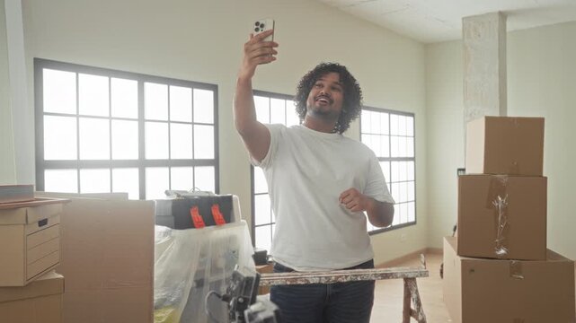 Man young indian in white tshirt holding smartphone, raised arm for selfie among packed boxes in building interior with ladder and toolbox; joy celebration.