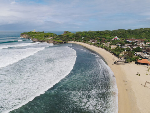 Aerial view of Baron Beach with turquoise waves crashing on the sandy shore, lush green cliffs, and coastal buildings in Tanjungsari, Special Region of Yogyakarta, Indonesia.