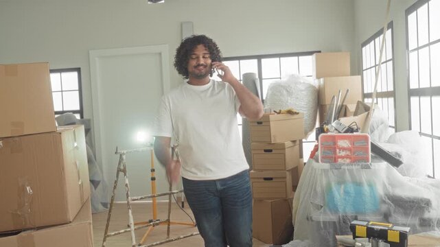 Young indian man holding smartphone to ear and resting hand on stacked moving boxes in a building filled with packing materials and tools; moving day relief.