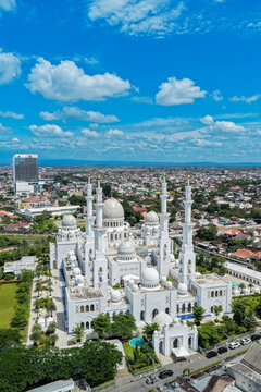 Aerial view of Sheikh Zayed Grand Mosque Solo Surakarta with its white domes and tall minarets under a bright blue sky with fluffy clouds in Kecamatan Banjarsari, Jawa Tengah, Indonesia.