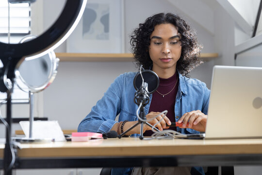 Non-binary adult adjusting microphone and handling small orange object at studio desk with laptop