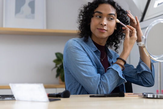 Non-binary person applying makeup with brush at home vanity using round mirror, wearing denim shirt