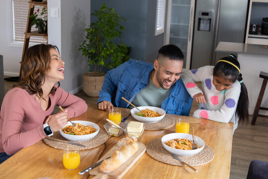 Diverse family sharing spaghetti at home kitchen table, laughing with daughter and orange juice