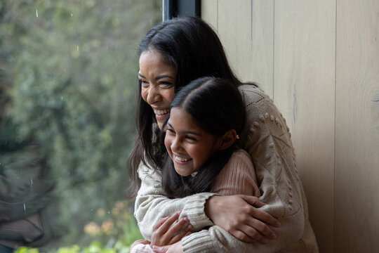 Indian mother and daughter sitting on window seat by rainy glass, wearing cream and pink sweaters