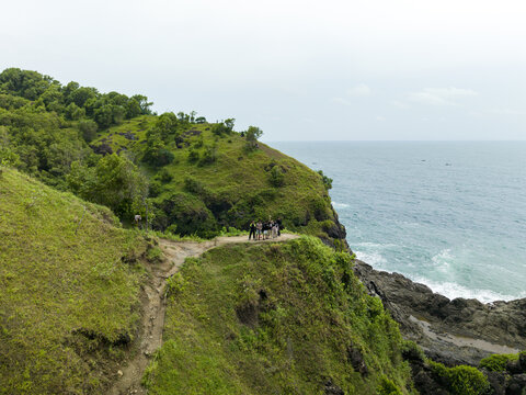 Aerial view of people standing on a coastal cliff edge near Banyutibo Waterfall with lush green hills and ocean waves in Girisubo, Special Region of Yogyakarta, Indonesia.